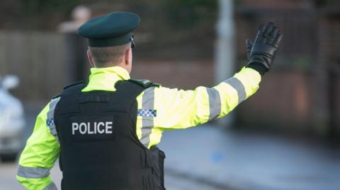 A PSNI officer is standing in the middle of a road wearing a protective vest, a green cap and a high-viz yellow jacket. His back is turned to the camera, has one hand on his hip and is holding the other out to flag down a car. 