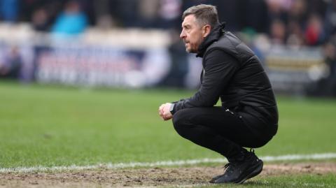 Port Vale boss Jon Brady is on his haunches on the touchline watching on during a game