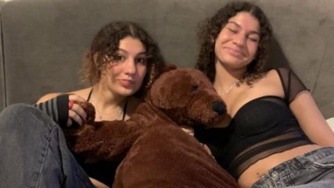 Twin sisters sit on a sofa with a dog cuddly toy between them