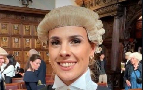 A female barrister in a wig smiles inside what appears to be a grand hall, with crowds of people behind her