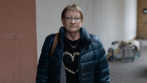 A woman with short hair and glasses stands with a grim expression wearing a dark coat. In the background, a shopping trolly can be seen tipped onto its side. 