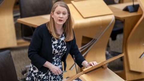 Shirley-Anne Somerville, wearing a black and white floral dress, stands at a podium in the Scottish Parliament chamber