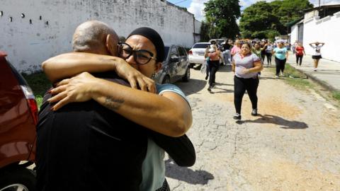 Two people are seen embracing tightly on a street in Tocuyito, Venezuela, with a group of people walking in the background and cars parked along the road.