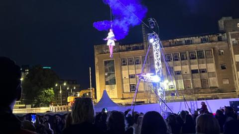 An acrobat wearing white is held by a wire high above a crowd of people at night in a city centre, with a derelict building in the background.