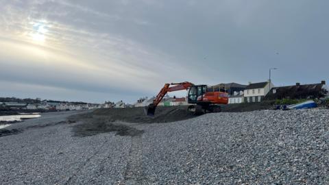 A digger on a rocky beach. There are houses in the background. The sky is grey and the sun is seen through the clouds. 