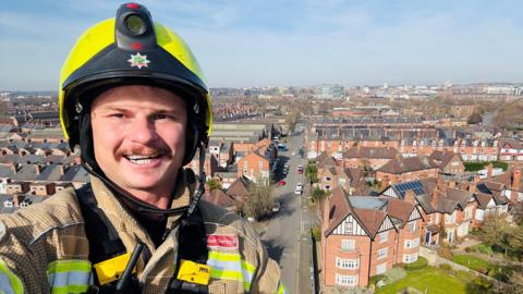 Tomasz Wisniewski in a firefighting uniform up on a ladder with homes in the background