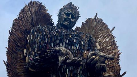 A tall male figure towers above a grey. The statues hands are open and shoulders are shrugged. The statue is made out of rusty knife blades of all different shapes and sizes.