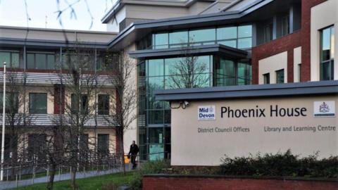 Exterior of Mid Devon District Council Phoenix House. The building is beige with red bricks and large windows. A man is walking out of the building. A pathway is lined with trees. 