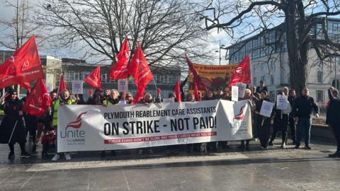 Carers lining up at a picket line in Plymouth, some are holding red banners which say union name Unite on them, they are holding a long rectangular banner which says Plymouth Reablement Care Assistants on strike - not paid! 