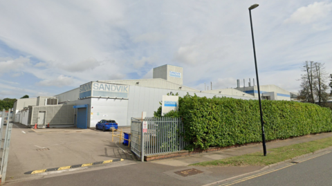 A street view of a large grey factory with blue signs and a hedge in front.