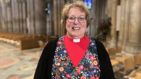 Rev Canon Fiona Brampton stands in Ely Cathedral facing the camera and smiling broadly. She is wearing a bright red shirt with a white dog collar, and over that a flowery patterned top and then a black cardigan. She has green glasses and short light brown curly hair. Chairs and pews are in the background, not in focus. 