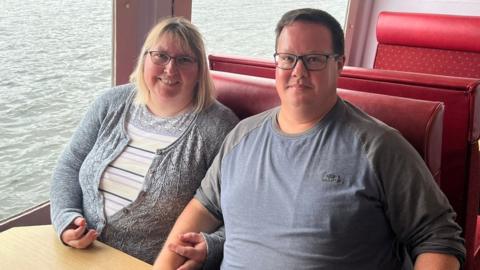 Sarah Luxton and Will Luxton sitting together on a red sofas next to water. She has linked her arm through his and they are both smiling.