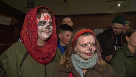 A man and woman, both wearing green coats with halloween face make upo