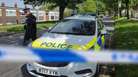 A police officer next to a police car behind blue and white police tape at a cordon set up after a World War Two bomb was found in Plymouth. The area is a residential street with houses on either side of the road. The police tape is blurred.