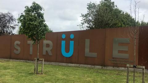 A brown wall with the word Strule written on it. There is grass and a tree in the forefront of the image. 