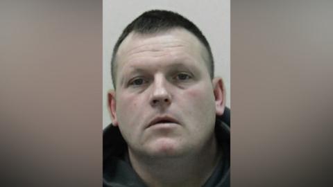Mugshot of David McNeil. He has pale pasty skin and a large clean shaven face with short black hair shaved at the sides