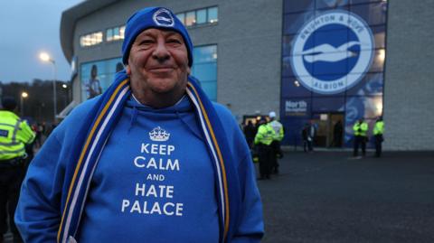 A man in a Brighton and Hove Albion beanie and hoodie that says "Keep calm and hate Palace". He is standing in front of a Brighton badge on the outside of the Amex.