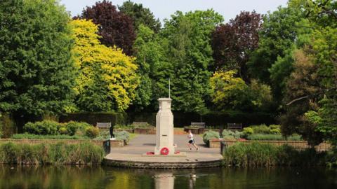 A war memorial stands by a pond surrounded by mature trees. A man is jogging past the memorial.