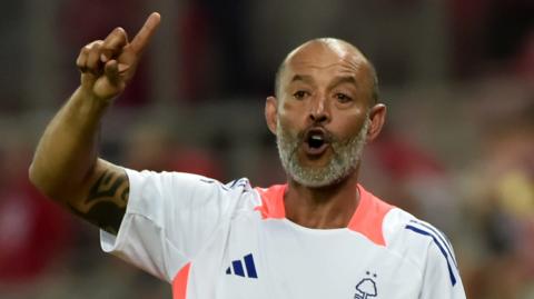 Nuno Espirito Santo holds his right arm in the air, raising his index finger, and opens his mouth while wearing a white Nottingham Forest t-shirt