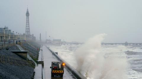 The spray of large waves rise as they crash into the coast of Blackpool with Blackpool Tower rising up behind on the left on a grey day. A maintenance vehicle is driven on a road next to the waves. The pier with a ferris wheel can be seen stretching over the water behind.