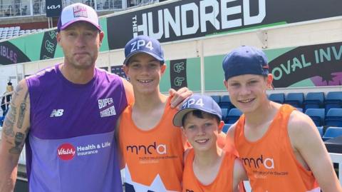 A man wearing a purple vest top and a white and purple hat poses for a picture with three boys all wearing orange vests and navy blue hats