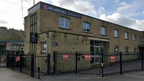 A yellow stone factory surrounded by black iron gates with a 'Millers Oils' sign
