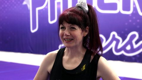 Imogen Ratcliffe is standing in front of a purple banner smiling and looking just off camera. She has her dyed red hair tied up in a high ponytail and is wearing a black vest.