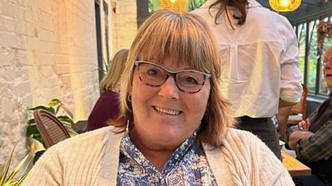 A woman sitting at a table in a restaurant. She has short blonde hair and is wearing tortoise shell glasses. She had a cream cardigan on a blue and white floral shirt. She is smiling and a waitress is standing behind the woman with her back to the camera.