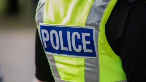 A police officer's torso seen from the back, they are wearing a short sleeved high viz vest with the word police in capital letters on a blue background. 