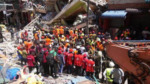 A large group of rescuers stands next to a huge pile of concrete rubble, while part of a crane can be seen in the foreground