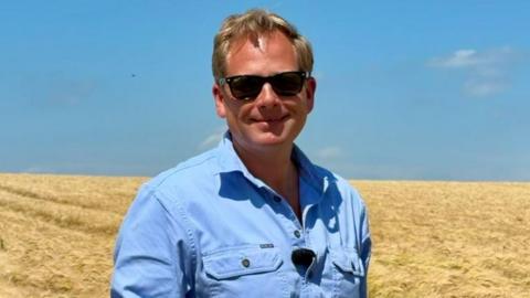 James Peck has short blond hair and is wearing black sunglasses and a blue shirt. He is standing in a field of yellow crops, and the sky is blue.