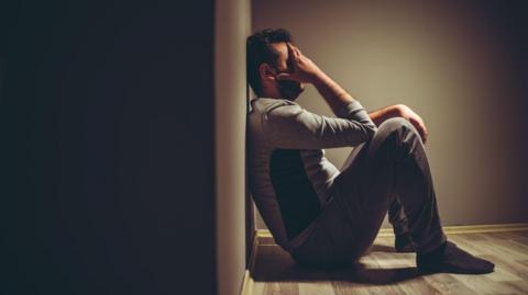 A stock image of man looking sad, sitting on the floor with his back against a wall