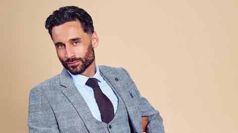 Model Luke Harden with black hair and beard wears a grey suit pale blue shirt and brown tie sitting on a chair in front of a beige background.