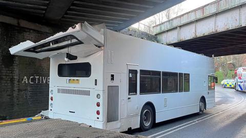 White double decker bus with roof ripped off after it hit a low bridge. A police and fire engine is seen past the bridge on a bend further up the street.