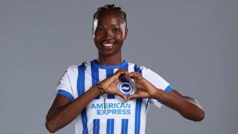 Aisha Masaka makes a heart shape over the Brighton crest while wearing a blue and white striped shirt