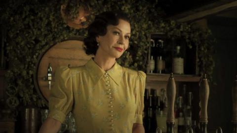 Catherine Zeta Jones in a yellow dress. She has short dark hair and is standing behind an old style bar. 