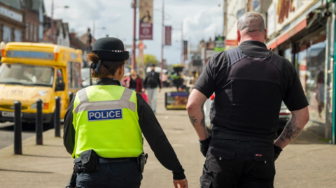 A police woman is walking alongside a security guard. The police officer wears a hat and her hair in a black claw clip while the security guard is bald but has tattoos on his head, and both forearms. He wears a black t-shirt and black body vest.