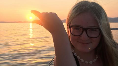 Lucy is wearing glasses and a necklace, posing as if she is trying to pinch the sun at sunset