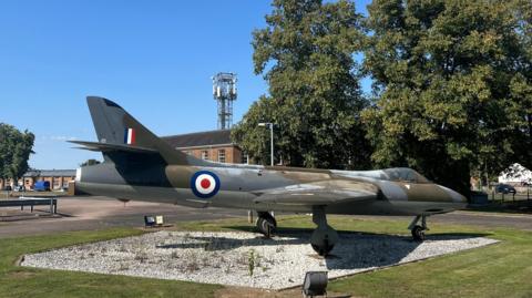 An RAF plane behind wire mesh fencing outside an airbase. The plane is a Hawker Hunter and has three wheels and a single cockpit.