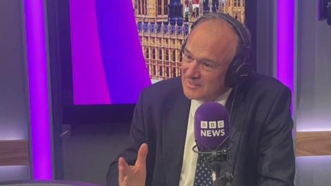 Sir Ed Davey in a BBC studio, wearing a white shirt, dark suit jacket and tie. He has headphones on and is talking into a purple BBC News microphone