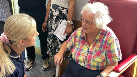Marjorie sits on a high backed red chair and wears a colourful checked shirt. She has got a walking stick in one hand. Katie Chalmers is in a nurse's uniform and kneels beside her to hear her talk. They are at Riverside Surgery in Bovey Tracey.