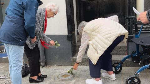 Two older women lay down flowers on a memorial stone. One if wearing a striped black and white jacket and red scarf, the other is wearing a cream jacket and a white floral top.