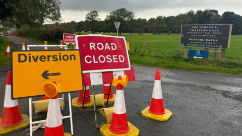 A road closure sign is at the heart of the picture with a diversion sign next to it and a number of traffic cones. A second road closure sign can be seen behind.