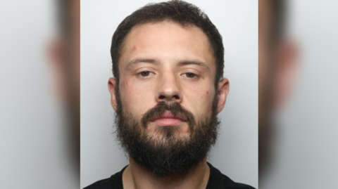 A mugshot of Daniel Kibblewhite. He wears a black t-shirt and has a dark brown beard and hair