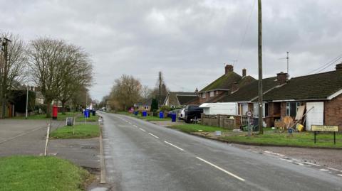 A general view of a street that through a village that is lined either side with houses. Blue rubbish bins sit close to the side of the road. Grass verges also separate the pavement from the road.