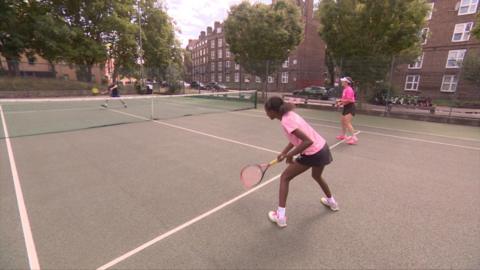 Young black girl wearing a pink top and black shorts on a tennis court ready to receive a ball
