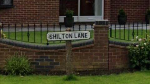 A road sign on the grass which reads Shilton Lane