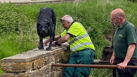 Two men removing the dog sculpture from the top of the wall next to the weir. Both are wearing green and one is wearing a hi-vis vest.
