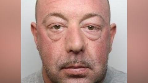 A close crop of a mugshot. A man with a pink face and short brown and grey stubble looks into the lens.