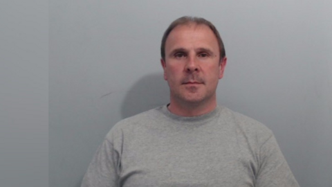 A mug shot of Irvine. It is a head and shoulders image, he is wearing a grey sweatshirt and has short, balding brown hair. He is looking directly down the lends of the camera.